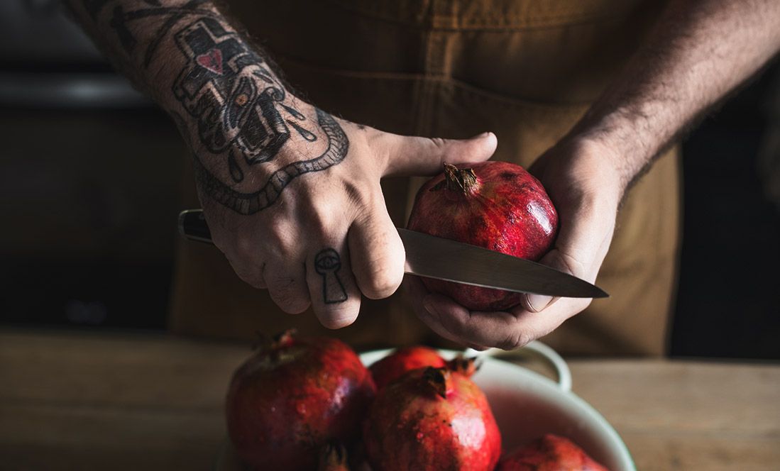 Hands of tattooed man cutting into a pomegranate over pomegranate bowl